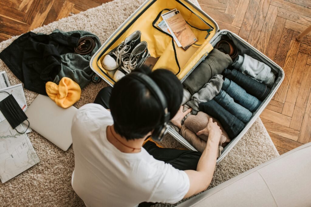 5 Best Travel Accessories That Actually Earned a Spot in My Bag Adult packing clothes in a suitcase while listening to music at home. Overhead view on rug.