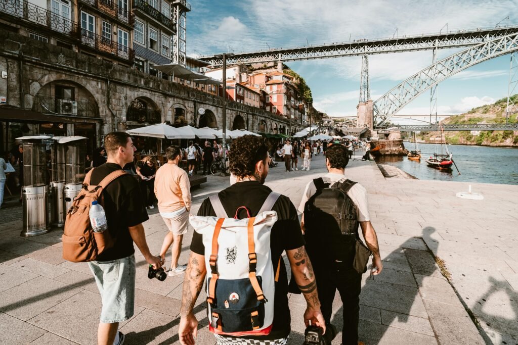 Free Walking Tours: The First Thing You Should Do In Any City Tourists with backpacks walk along Porto's scenic riverside promenade under a sunny sky.