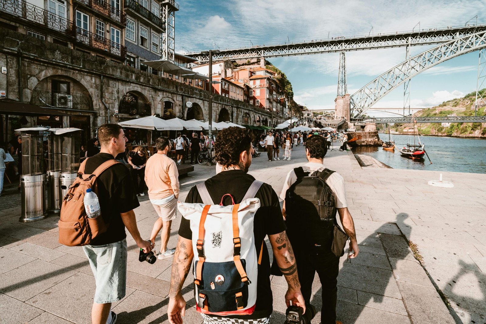 Free Walking Tours: The First Thing You Should Do In Any City Tourists with backpacks walk along Porto's scenic riverside promenade under a sunny sky.